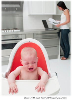 An infant is in a high chair crying while the mother appears to be looking at paperwork or bills at a computer.