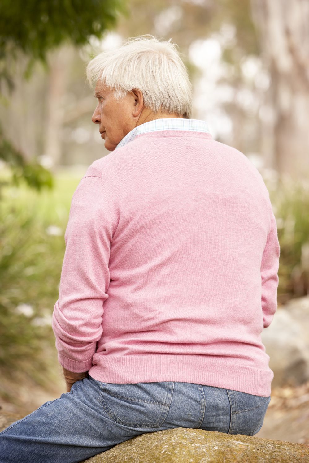 Elderly person in sweater and jeans sitting on rock, with back to camera.