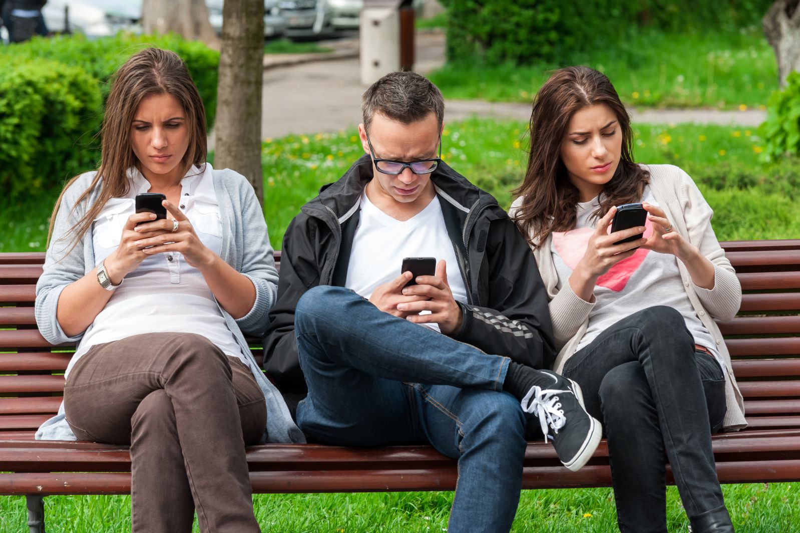 Color image of two female teens and one male teen sitting on park bench, staring at their smartphones.