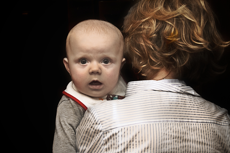 Photo shows an infant age 6-12 months hugging a parent. The infant’s expression is confused and astonished. The parent’s face does not visible.