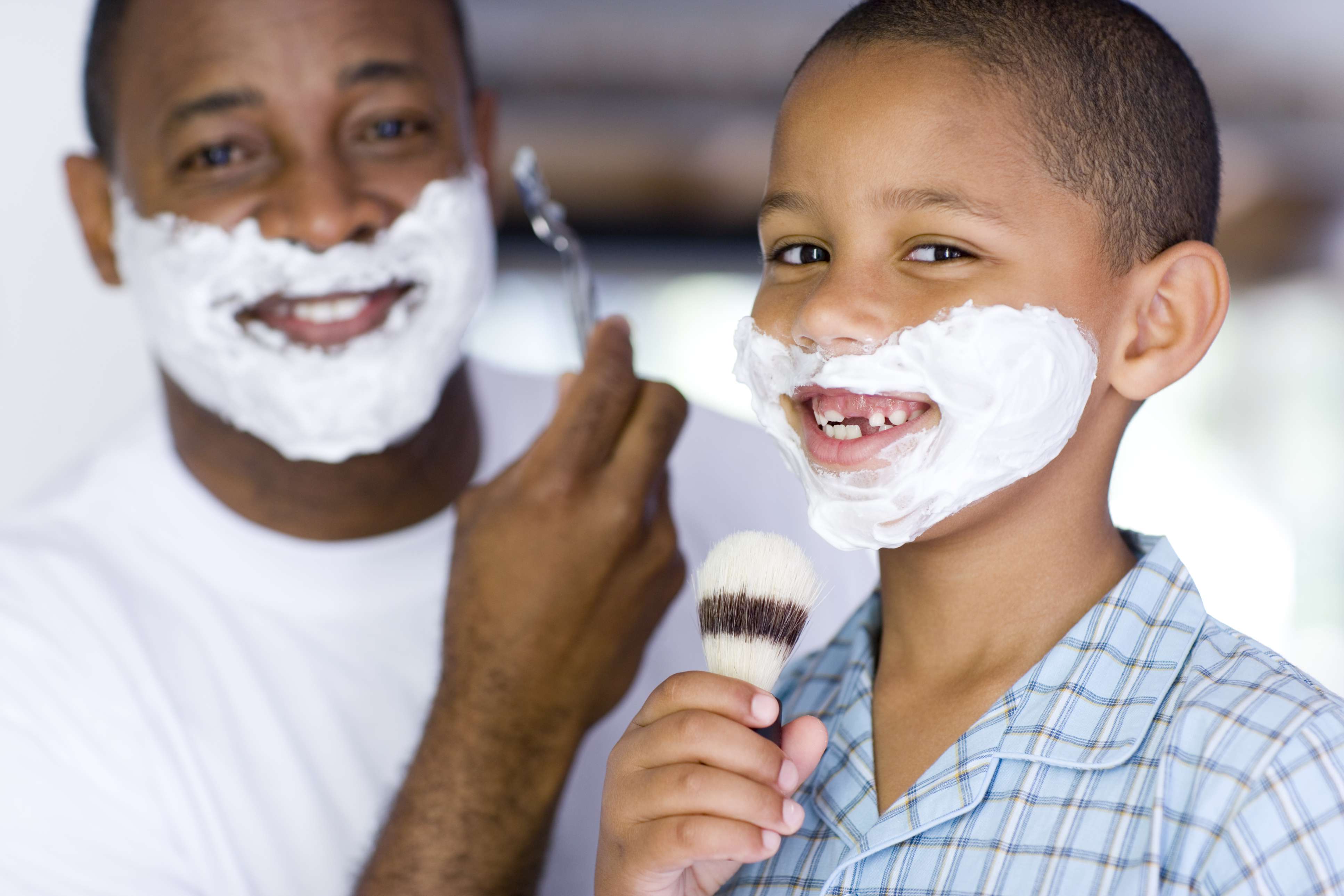The image shows a boy and his father. Both have shaving cream on their face. The man has a razor in his hand, while the boy holds a shaving cream brush.