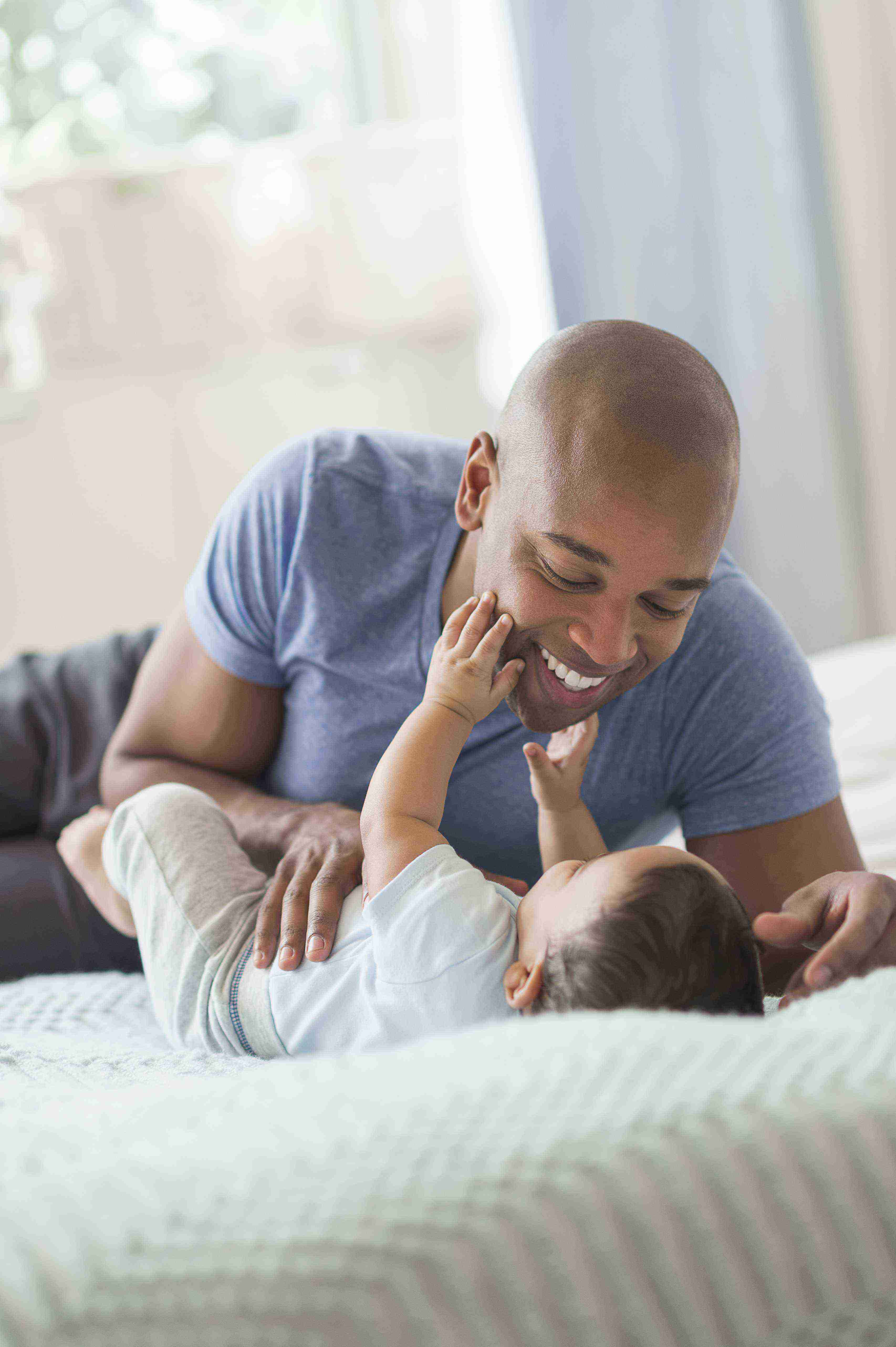 A baby is laying on a bed touching the face of a smiling man leaning over him