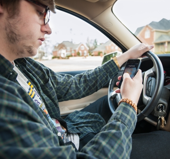 A young man looks at his cell phone as he drives his car.