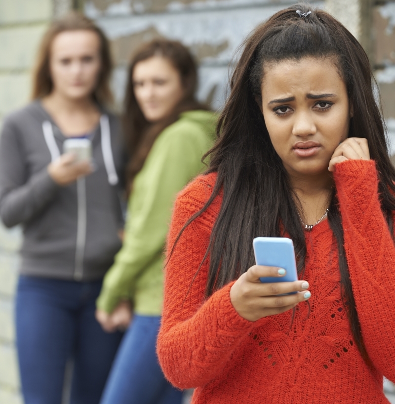 A teenage girl dressed casually in a crimson-colored sweater looks upset as she reads something on her cell phone and two young women glance at her from behind.