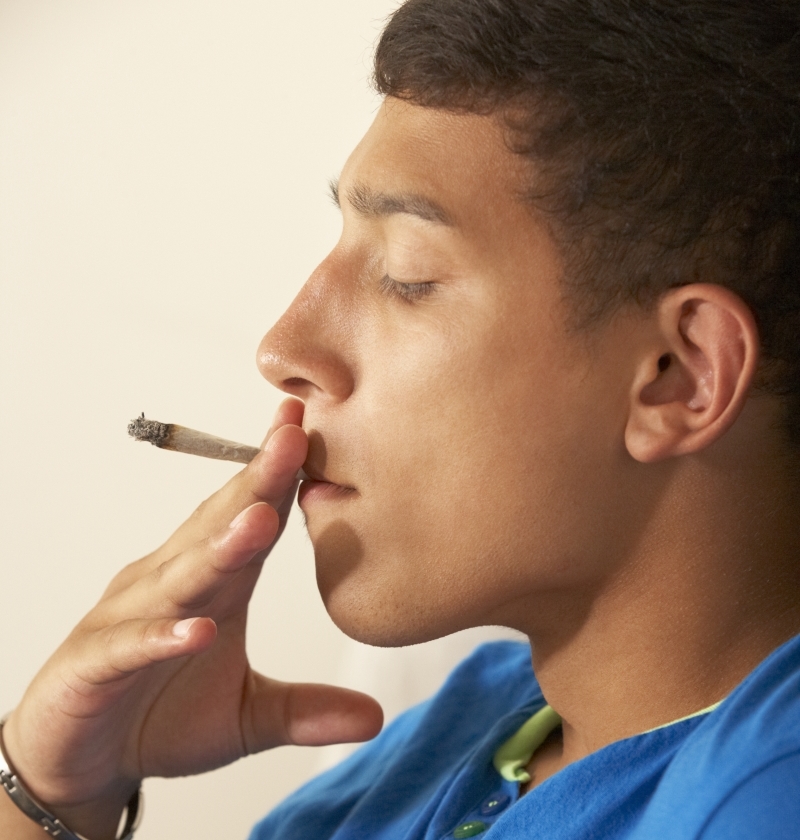 A teenager in a blue colored t-shirt  holds a cigarette to his lips.