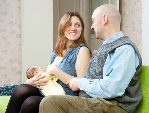 A lady breastfeeds her baby and smiles at her partner who sits next to her.