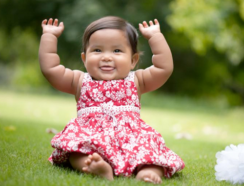 A baby girl in a red floral dress smiles with her hands raised up.