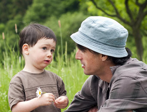 A toddler holds a flower and stands next to his father in a green meadow.