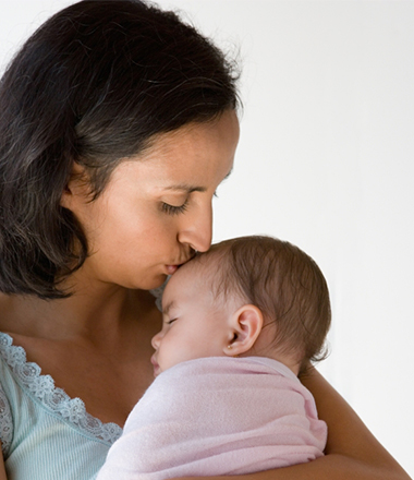 A mother holds her newborn baby close to her chest, kissing the baby on the forehead. The baby is wrapped in a pink towel and the eyes are closed.