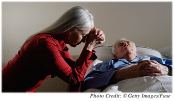 An older woman praying at the bedside of her ailing spouse 