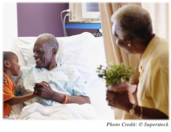 An older man in a hospital bed receiving a visit from a grandchild or great grandchild and a spouse