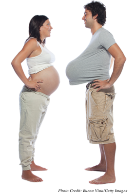 A pregnant woman with her stomach exposed facing a man with a balloon under his shirt that looks like a pregnant belly.  They are smiling at each other.