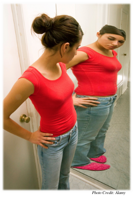 A girl looking at herself in a full length mirror.  She is of a healthy-looking body weight, yet she sees her reflection as significantly heavier than her actual body.