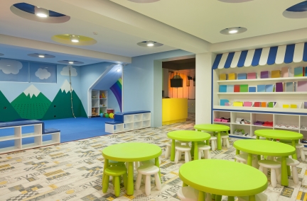  a classroom with one side having green tables and stools on tile floor with various colored boxes on a wall.  The other side of the room has a mountain range painted on the wall, and has blue carpet.