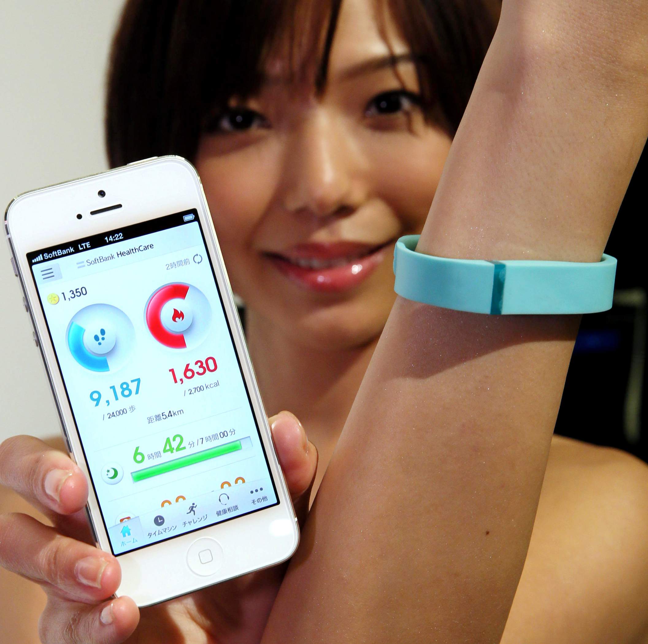 A young woman wears a wearable activity tracker and shows her smart phone tracking the fitness data from the tracker on her wrist.