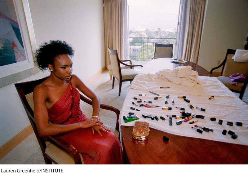 A photo of a woman sitting at a table strewn with lipsticks and other items of makeup. The woman has a dazed look on her face and is not looking directly at the table.