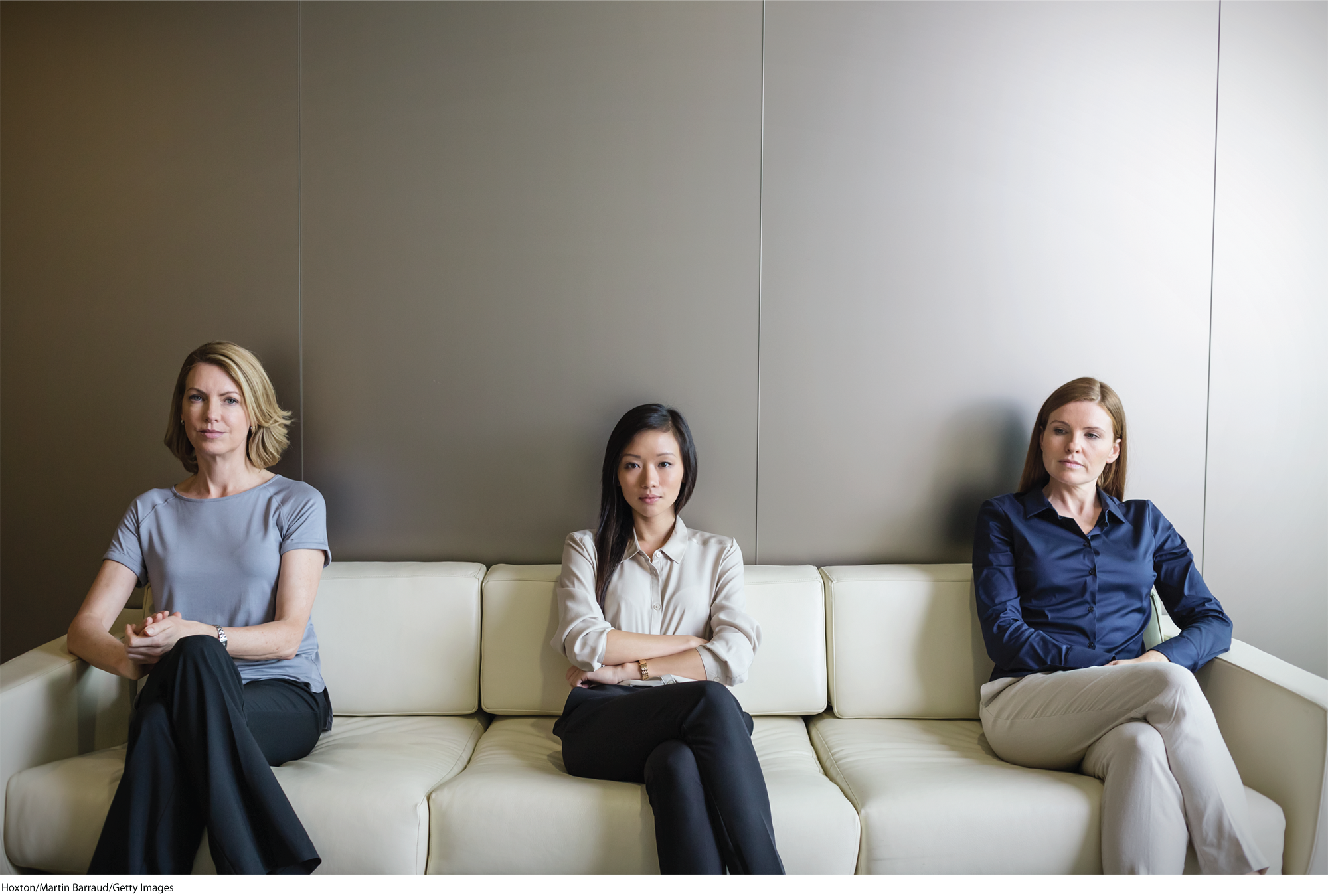 A photo shows three young women sitting on a couch. They are sitting at a distance from each other.