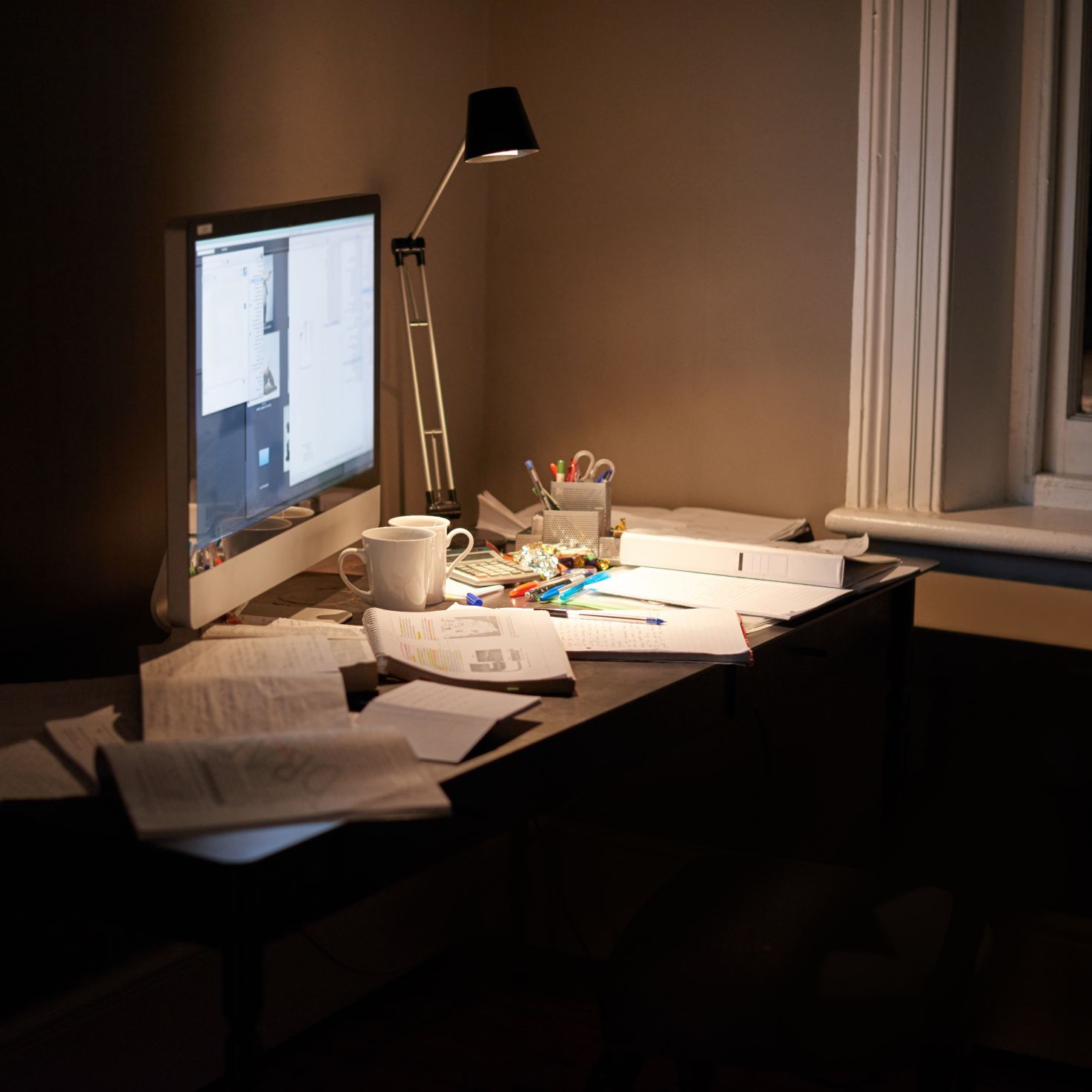 Scattered papers and textbooks on a desk with computer screen and lamp.