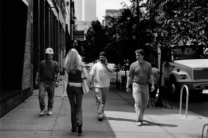 Color photo of a blonde woman walking past three men on a city street. The photo is taken from behind the woman as the three men walk toward the camera, showing the facial expressions of the men. The men are all smiling and looking at the woman as they walk past. The woman’s face is not visible.