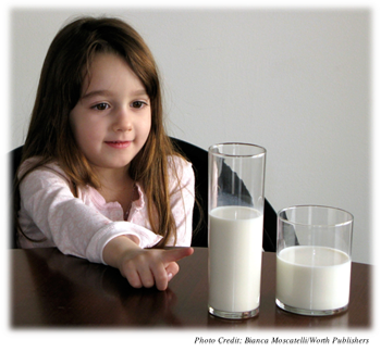 A preoperational age girl studying two different shaped glass filled with the same amount of liquid.  Because they are different shapes, the girl is pointing to to the taller, thinner one because it appears to have more liquid.