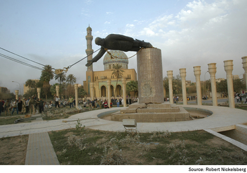 Images. A photograph of the same scene taken from a distance. It shows the statue being lowered from the column in the middle of the frame and an empty pavilion in front of the column and statue. In the background is a group of people, at some distance from the column and statue. Source Robert Nickelsberg.