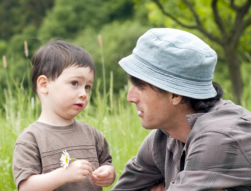 A toddler holds a flower and stands next to his father in a green meadow.