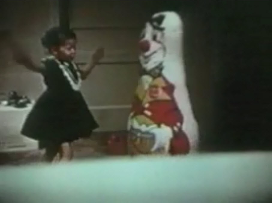The image is a still photo of a girl walking towards a Bobo doll with her hands raised in the air.