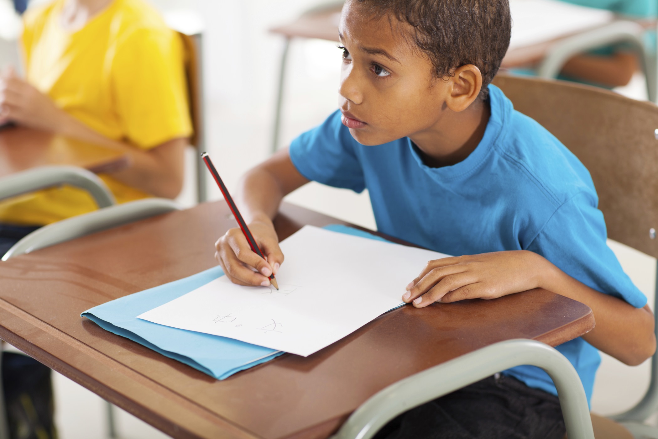 An older boy is sitting at a desk in a classoom writing on a paper with a pencil. He is between 7 and 11 years old