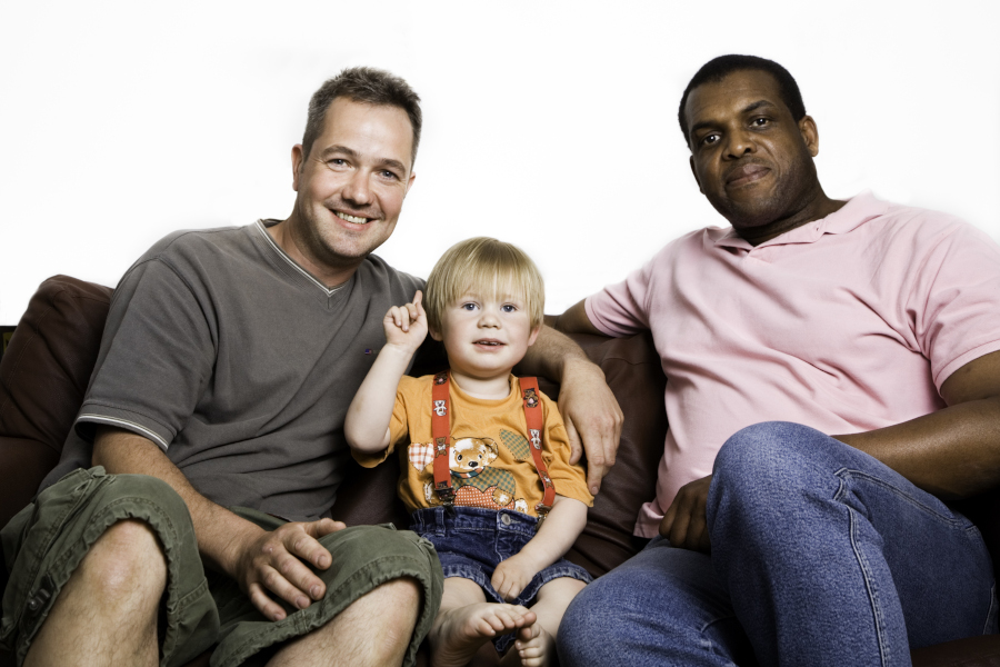 Unconventional multiracial family: twomen, black and white, with a boy sitting on sofasmiling, looking in camera.
