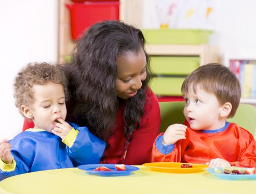 Two kids eat fruits in day care as the female care taker watches them. 