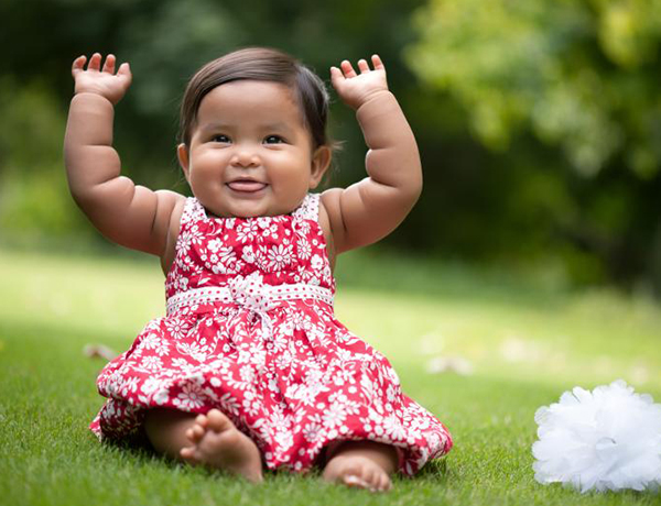 A baby girl in a red floral dress smiles with her hands raised up.