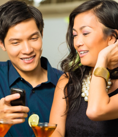 A young, well-dressed Asian couple look at a cell phone message together while enjoying a cocktail.