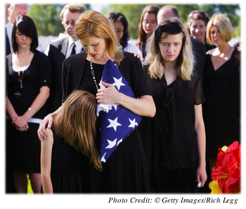 Mourners of all ages dressed in black at a military gravesite.  The spouse  of the deceased is comforting her daughter with one arm and holding the US flag  folded up in the other arm.