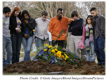 High school-aged mourners, some holding flowers, gathered around a new gravesite