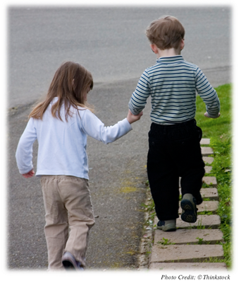 An older sister holding her brother's hand as he walks along balancing on a curb
