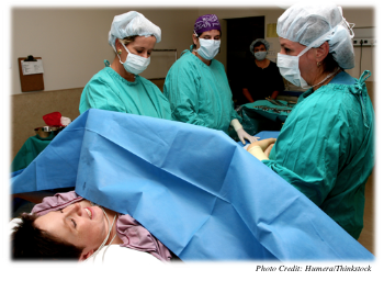 A mother-to-be is lying in an operating room with a sheet blocking her view of her abdomen.  Doctors and nurses are in scrubs prepared to perform a cesarean section.