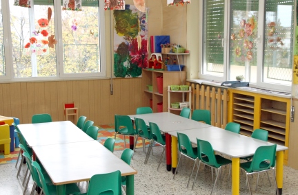 A classroom with two long white tables with green chairs.  There is some artwork hanging from the ceiling.