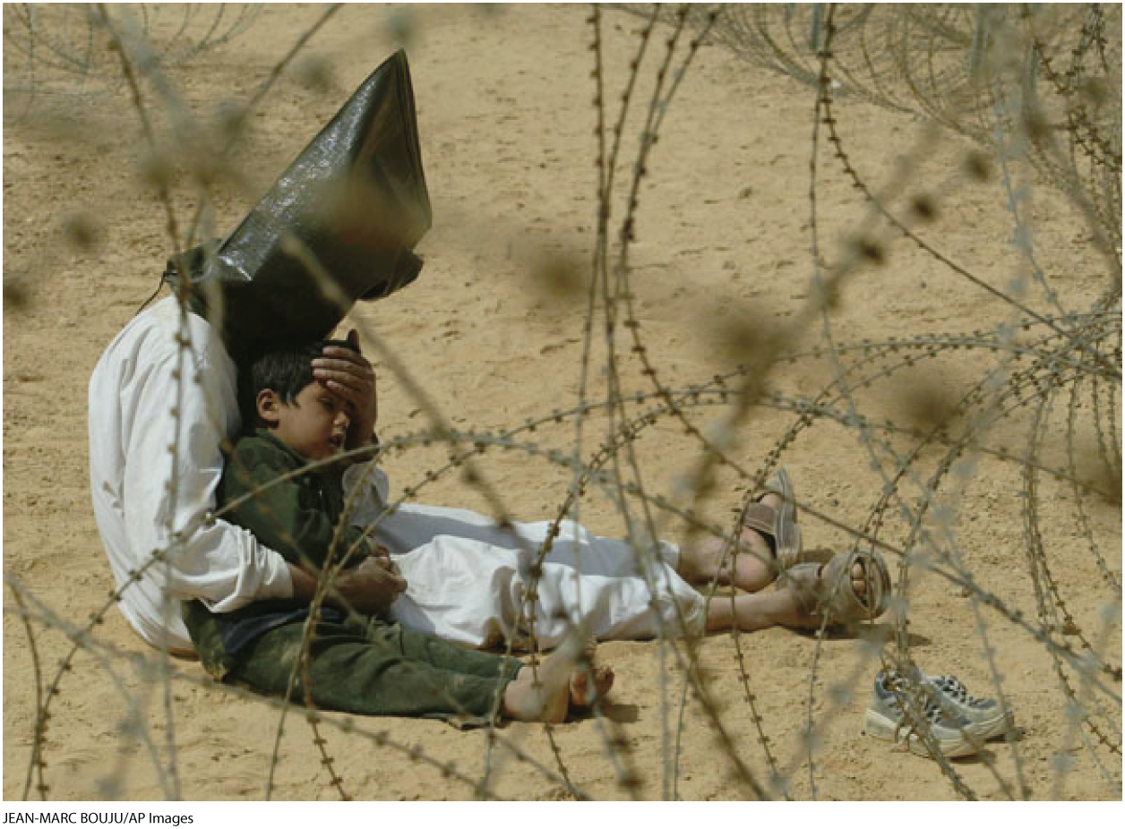 A photo shows a man sitting behind a barbed wire fence with a small boy. Both of them are sitting on a sandy ground. The man is holding the child close to him and has placed his hand over the child’s forehead as to comfort him. The child is barefoot and his shoes are shown at a distance. The man is wearing a head bag. A button on the left corner reads “Enlarge” and shows a plus icon.