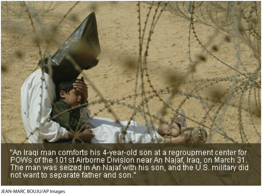 A photo shows a man sitting behind a barbed wire fence with a small boy. Both of them are sitting on a sandy ground. The man is holding the child close to him and has placed his hand over the child’s forehead as to comfort him. The child is barefoot and his shoes are shown at a distance. The man is wearing a head bag. The text below reads, An Iraqi man comforts his 4-year-old son at a regroupment center for POWs of the 101st Airborne Division near An Najaf, Iraq, on March 31. The man was seized in An Najaf with his son, and the U.S. military did not want to separate father and son.