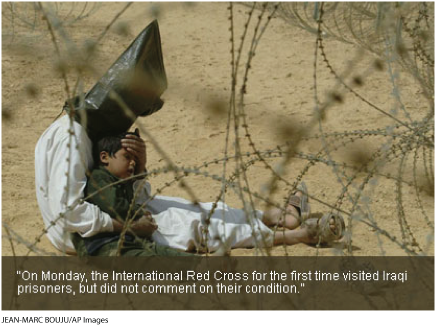 A photo shows a man sitting behind a barbed wire fence with a small boy. Both of them are sitting on a sandy ground. The man is holding the child close to him and has placed his hand over the child’s forehead as to comfort him. The child is barefoot and his shoes are shown at a distance. The man is wearing a head bag. On the left corner a button shows Enlarge with a plus icon. The text below reads, On Monday, the international Red Cross for the first time visited Iraqi prisoners, but did not comment on their condition.
