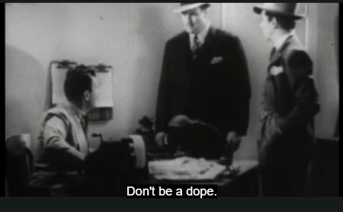 One of the men is sitting near a table and two men wearing hats are standing. The man who is sitting is looking at the man standing next to him. The subtitle reads Don't be a dope.