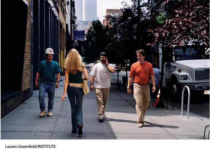Color photo of a blonde woman walking past three men on a city street. The photo is taken from behind the woman as the three men walk toward the camera, showing the facial expressions of the men. The men are all smiling and looking at the woman as they walk past. The woman’s face is not visible.