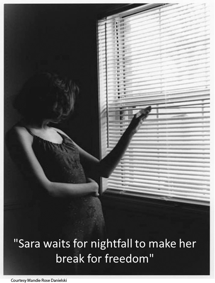 Black-and-white photo of woman peeking through window blinds. The title at the bottom reads, “Sara waits for nightfall to make her break for freedom.”