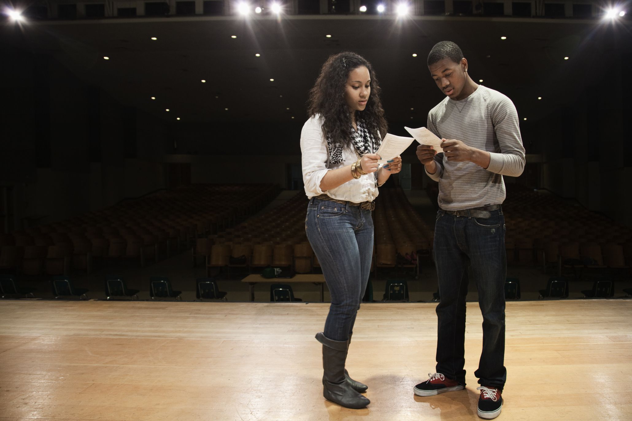 A teenage girl and boy stand on a stage reading their scripts. Stage lights and seats in the background.