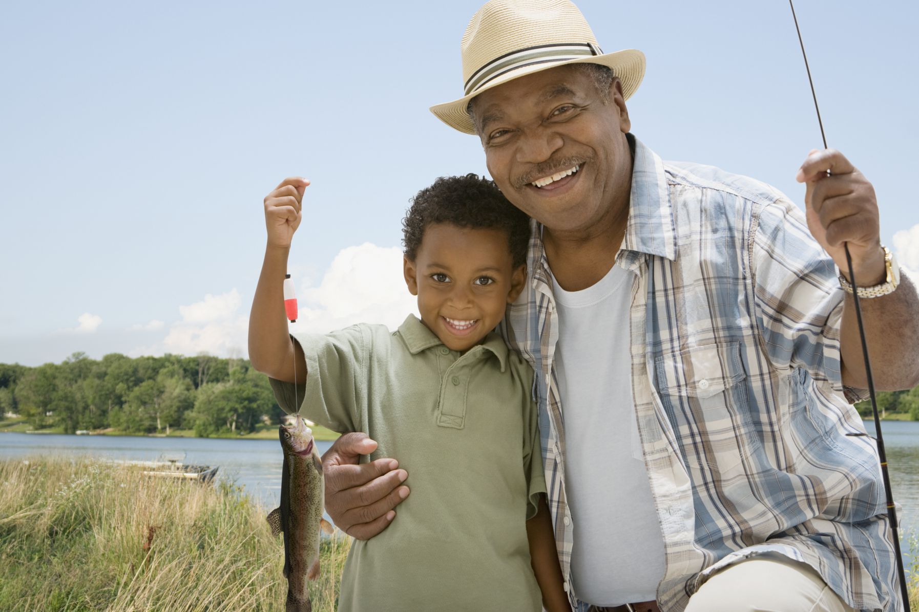 A boy holding a fish and his grandfather holding a fishing pole in front of a body of water.