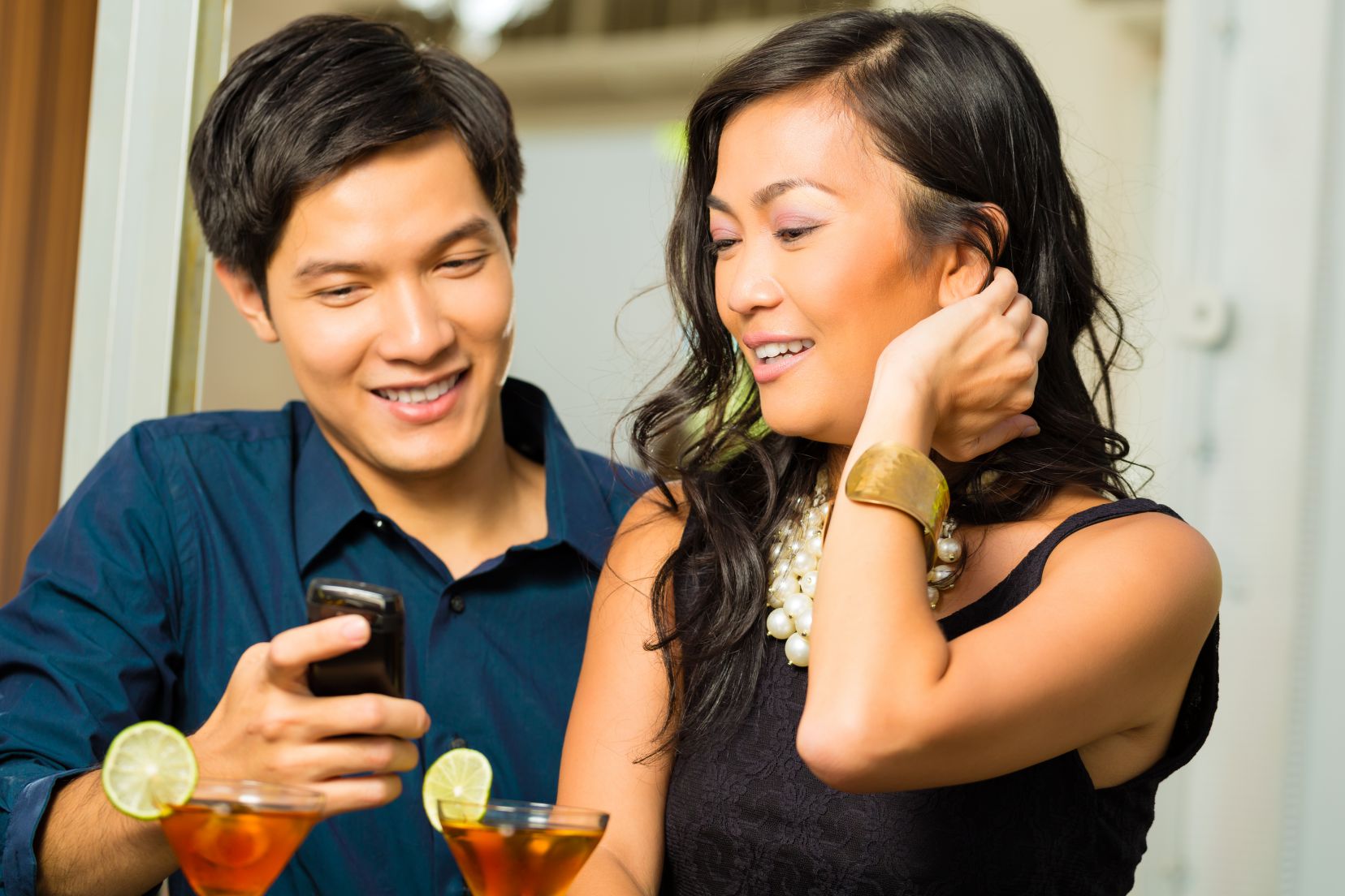 Asian man is flirting with woman in a bar while having drinks, woman is shy