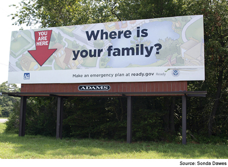 Image. Photograph of a large billboard. The billboard has a map of a small town in the background and the words You are here and a large arrow pointing to one house on the map. The largest words, in the center of the billboard, are Where is your family? Underneath in a smaller font are the words Make an emergency plan at ready.gov