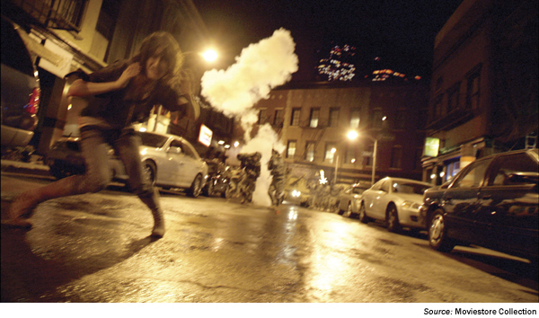 Image. Photo still from the movie Cloverfield. The image shows a street scene at night, with a distant spotlight providing illumination, a figure in the foreground, cars lining the street, and several figures and clouds of smoke in the background. The camera seems to be positioned on the ground in front of the figure in the foreground. Source Moviestore Collection.